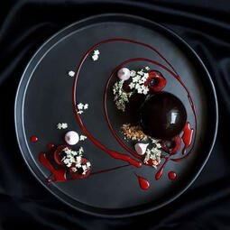 Dark Pearl Cake. A round dark chocolate cake with white chocolate garnish, flowers, and red jam, served on a black plate for a striking look.